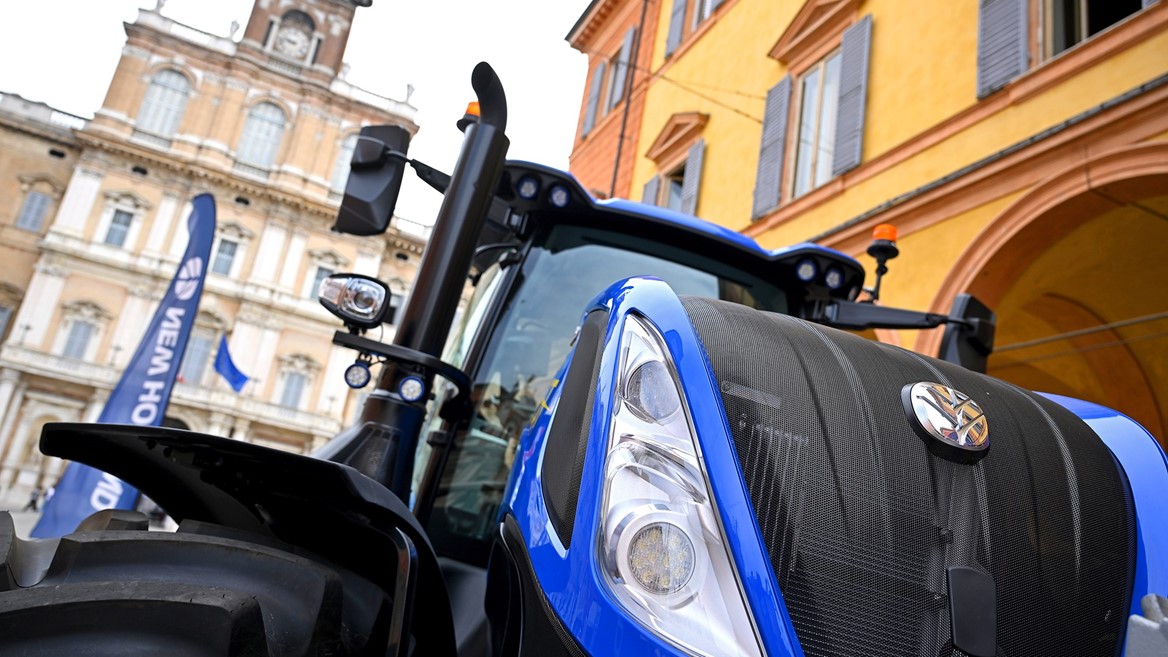 Close-up view of a blue New Holland tractor displayed outdoors in front of historic European-style buildings with arched windows and shutters, under a partly cloudy sky.