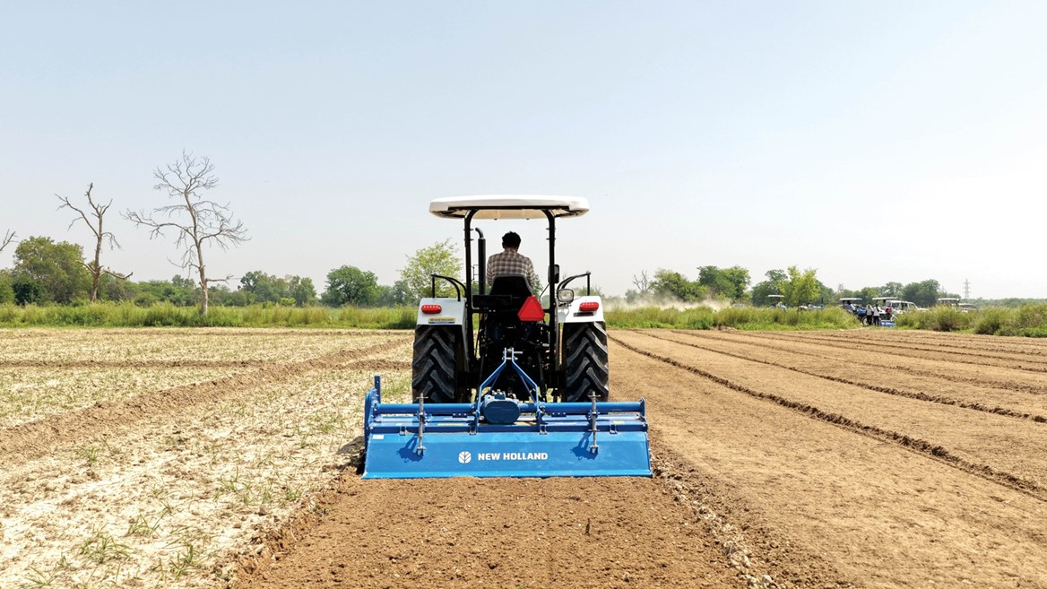 A farmer driving a white New Holland tractor with a blue tiller attachment, plowing a large, dry agricultural field under a clear sky, with green trees and other tractors visible in the background.
