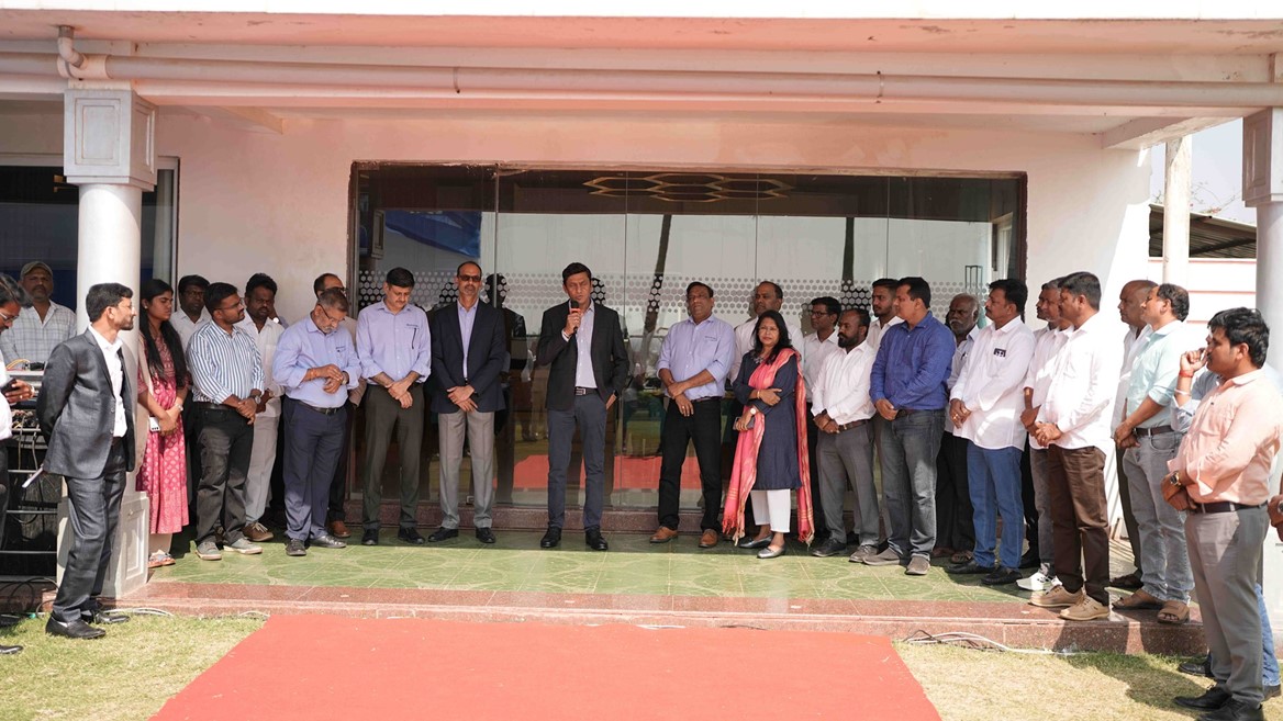 A group of professionally dressed men and women standing in a semi-circle outside a building with a red carpet laid out in front. One man in the center is speaking into a microphone, suggesting a formal event or gathering. The background shows glass doors and a white exterior wall.