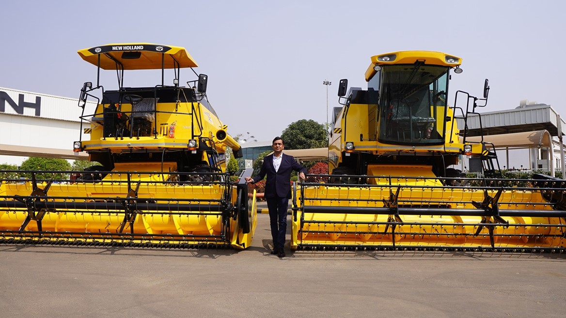 A man in a dark suit standing between two large yellow New Holland agricultural combine harvesters parked outdoors on a paved surface, with a building and greenery in the background under a clear sky.