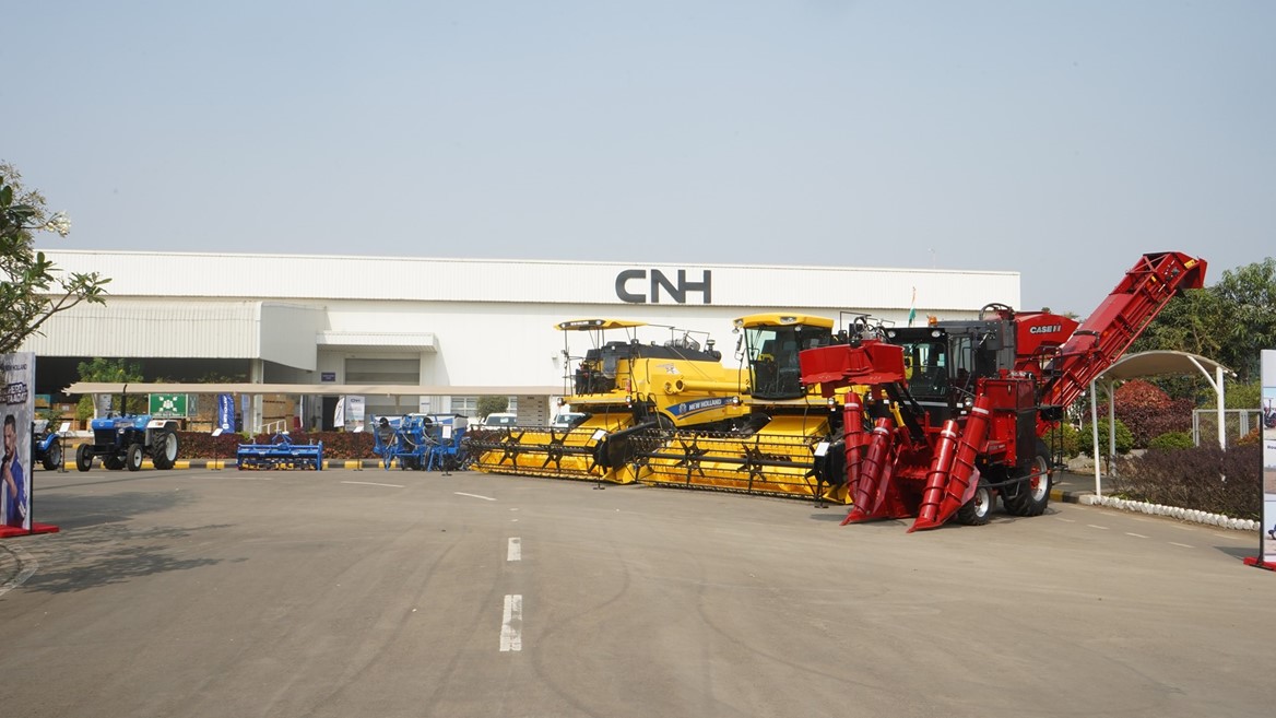 Outdoor display of large agricultural machinery including yellow combine harvesters and a red corn harvester parked in front of a white industrial building with the CNH logo, under a clear blue sky.