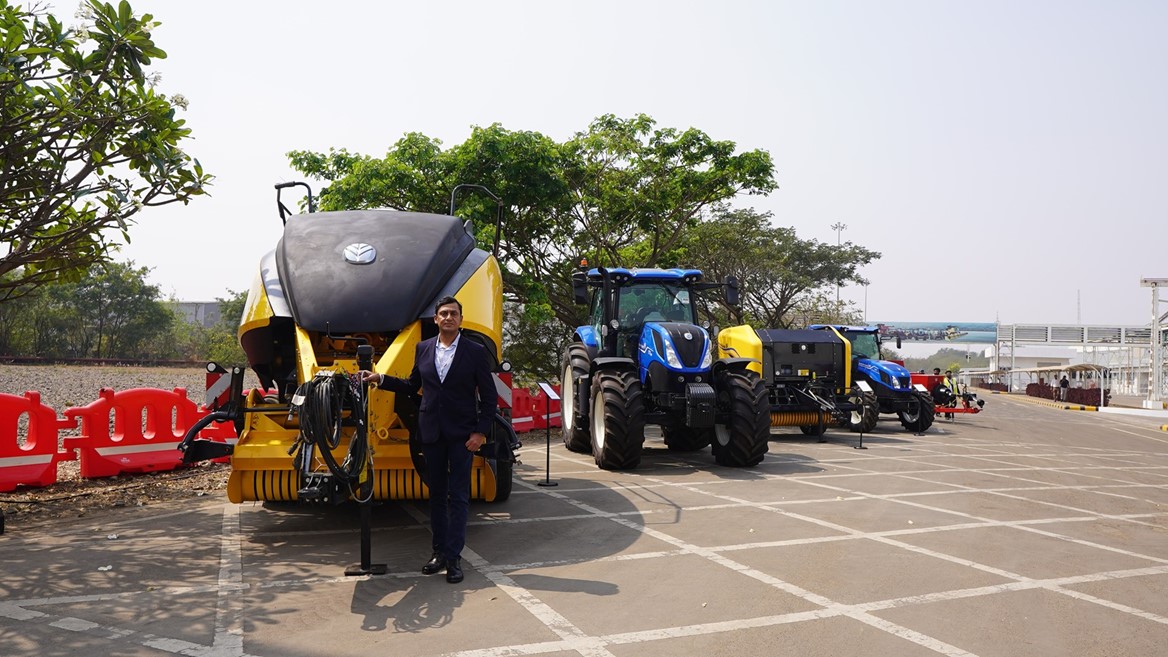 A man in a dark suit stands next to large yellow and black agricultural machinery and blue tractors parked in a row on a paved outdoor area with trees and red barriers in the background under a clear sky.