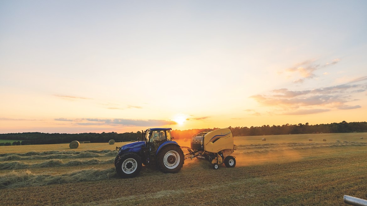 A blue tractor pulling a yellow baler machine in a large open field during sunset, with round hay bales scattered across the field and a clear sky with soft clouds in the background.