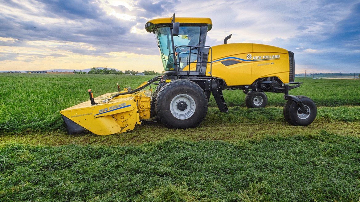 A yellow New Holland agricultural harvester machine is parked on a green field under a partly cloudy sky during daylight, with freshly cut crops visible on the ground.