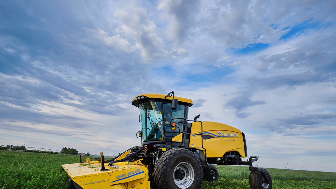 A yellow agricultural mower machine cutting green grass in a vast field under a partly cloudy blue sky during daytime.