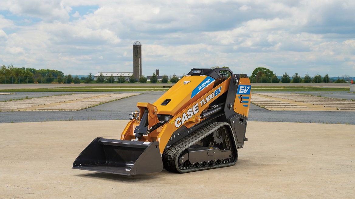 A yellow CASE TL100EV electric mini track loader with black tracks is parked on a concrete surface outdoors under a partly cloudy sky with a rural landscape and silo visible in the background