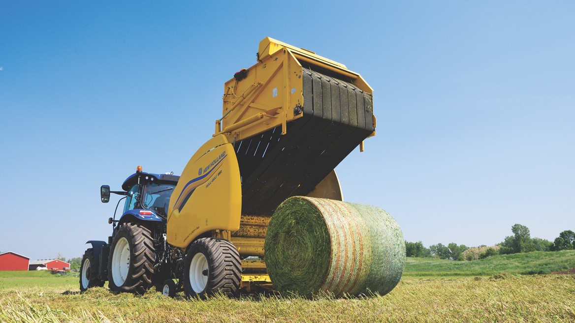 A blue tractor attached to a yellow New Holland round baler machine is operating in a green field under a clear blue sky, producing a large round bale of hay. In the background, there are some trees and a red barn.