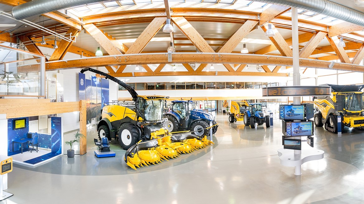Interno di un moderno showroom agricolo con macchinari agricoli di grandi dimensioni, tra cui trattori blu, mietitrebbie gialle e attrezzature per la raccolta, illuminato da ampie finestre e travi in legno a vista.