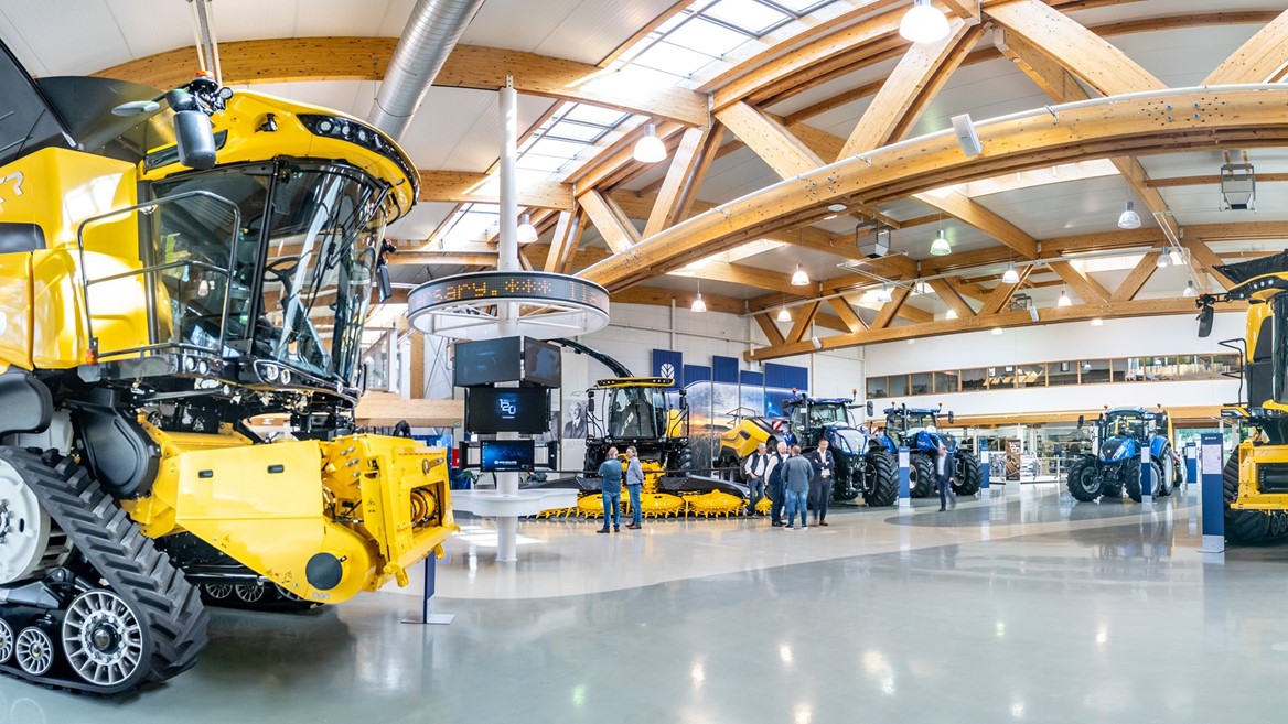 Interno di un grande showroom con macchinari agricoli moderni di colore giallo e blu, tra cui mietitrebbie e trattori, con alcune persone che osservano e discutono vicino ai veicoli, struttura con travi in legno e ampie finestre che lasciano entrare molta luce naturale.