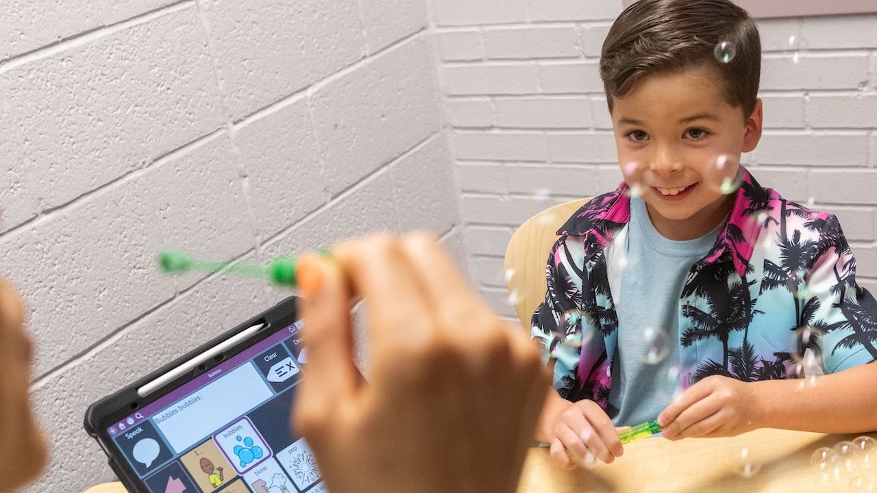 IMAGE DESCRIPTION A boy looks intently at a woman blowing bubbles while a tablet on a table displays learning icons