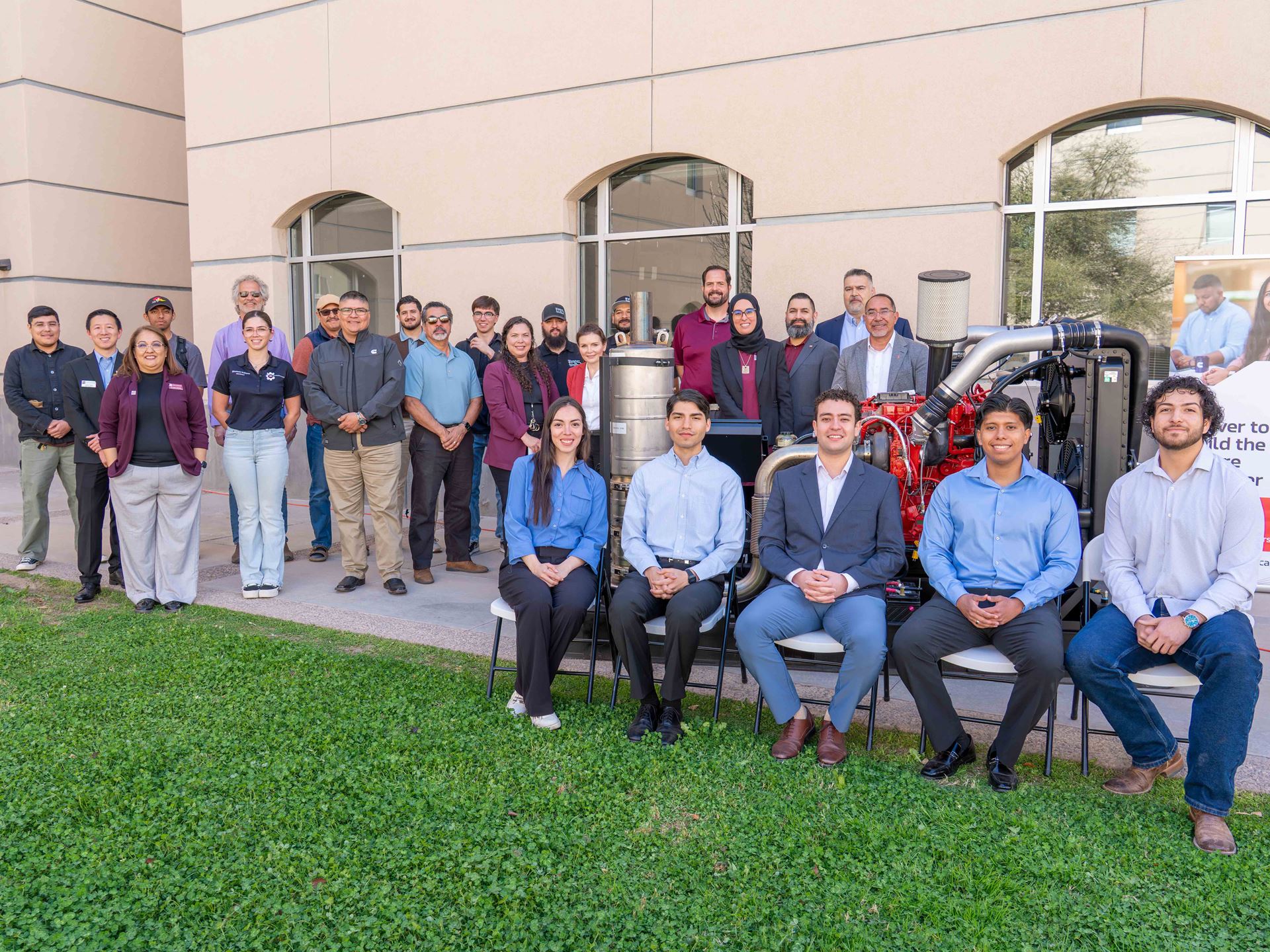 Description A group of people stand and sit next to and behind a large diesel engine that is displayed in a courtyard