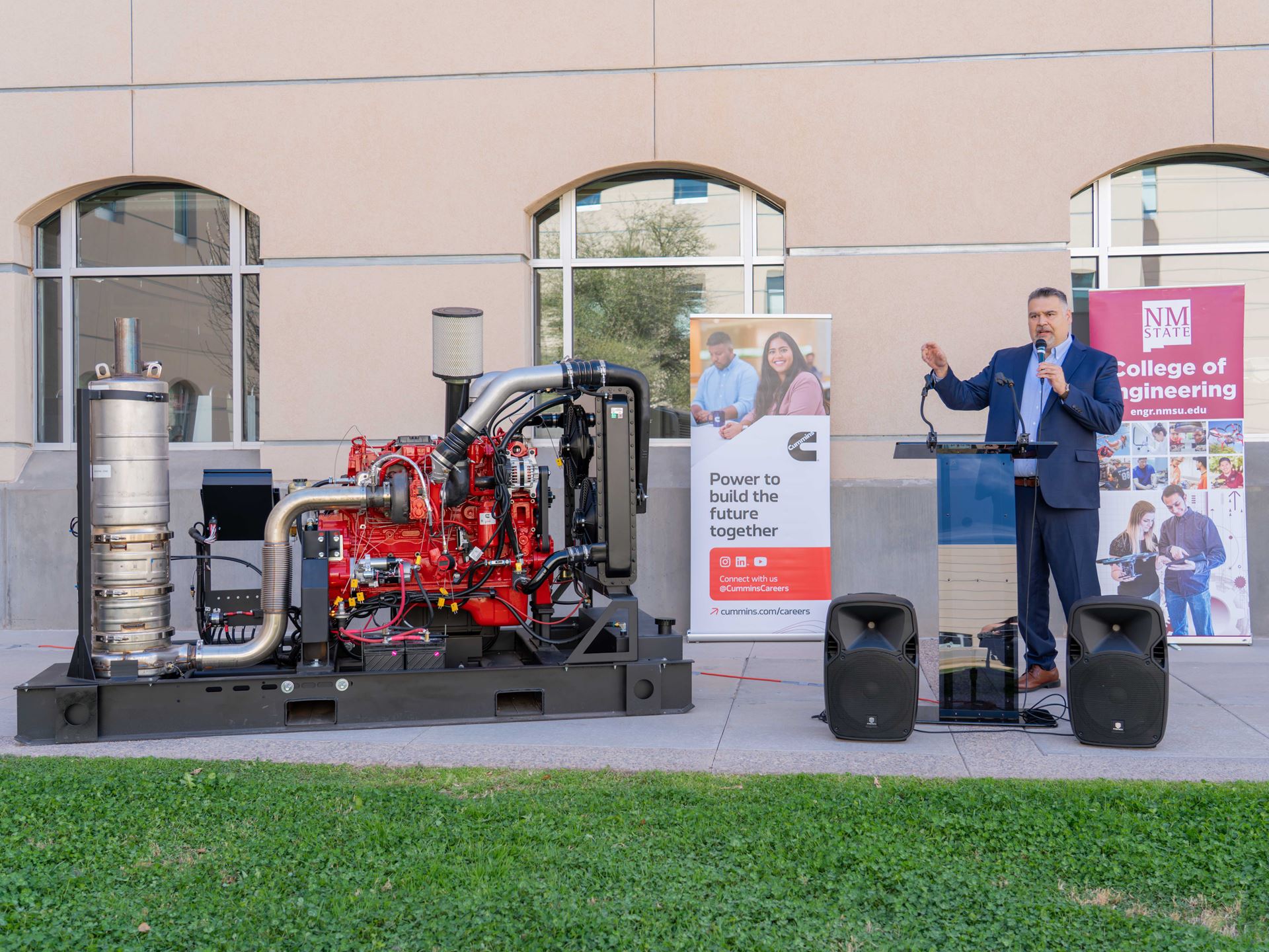 Description A man in a suit stands next to a diesel engine while speaking at an event