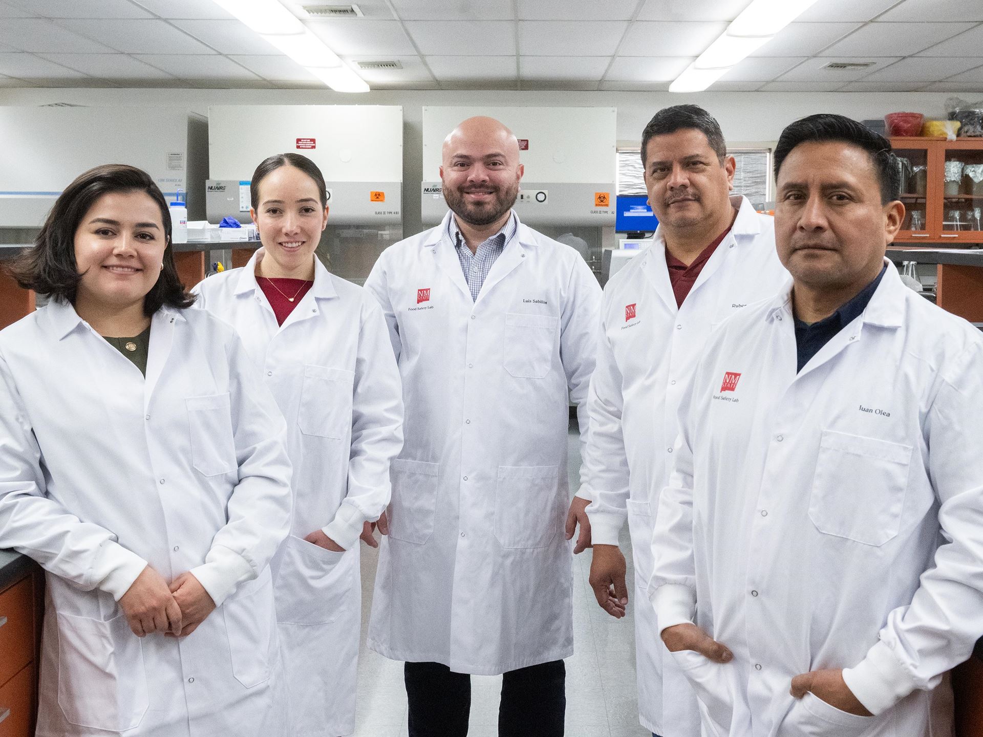 Two women and three man each wearing lab coats standing in the middle of a laboratory