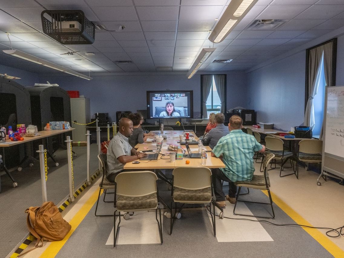 IMAGE DESCRIPTION A group of people seated around a table watching a woman speak on a monitor