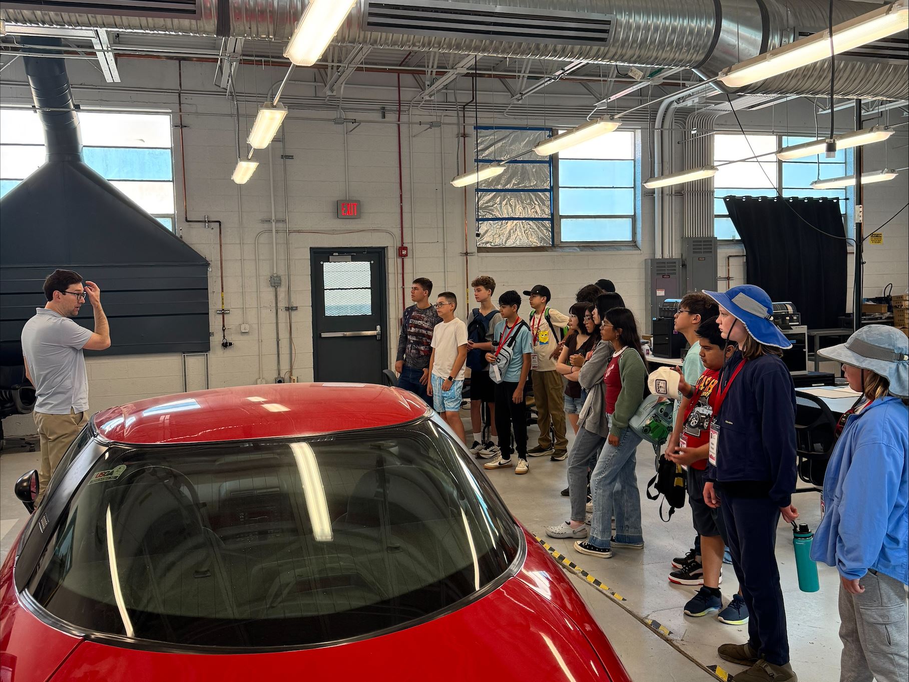 Description Middle school students attending NM PREP Academy are gathered around a red electric car in a mechanical engineering lab while they listen to an instructor