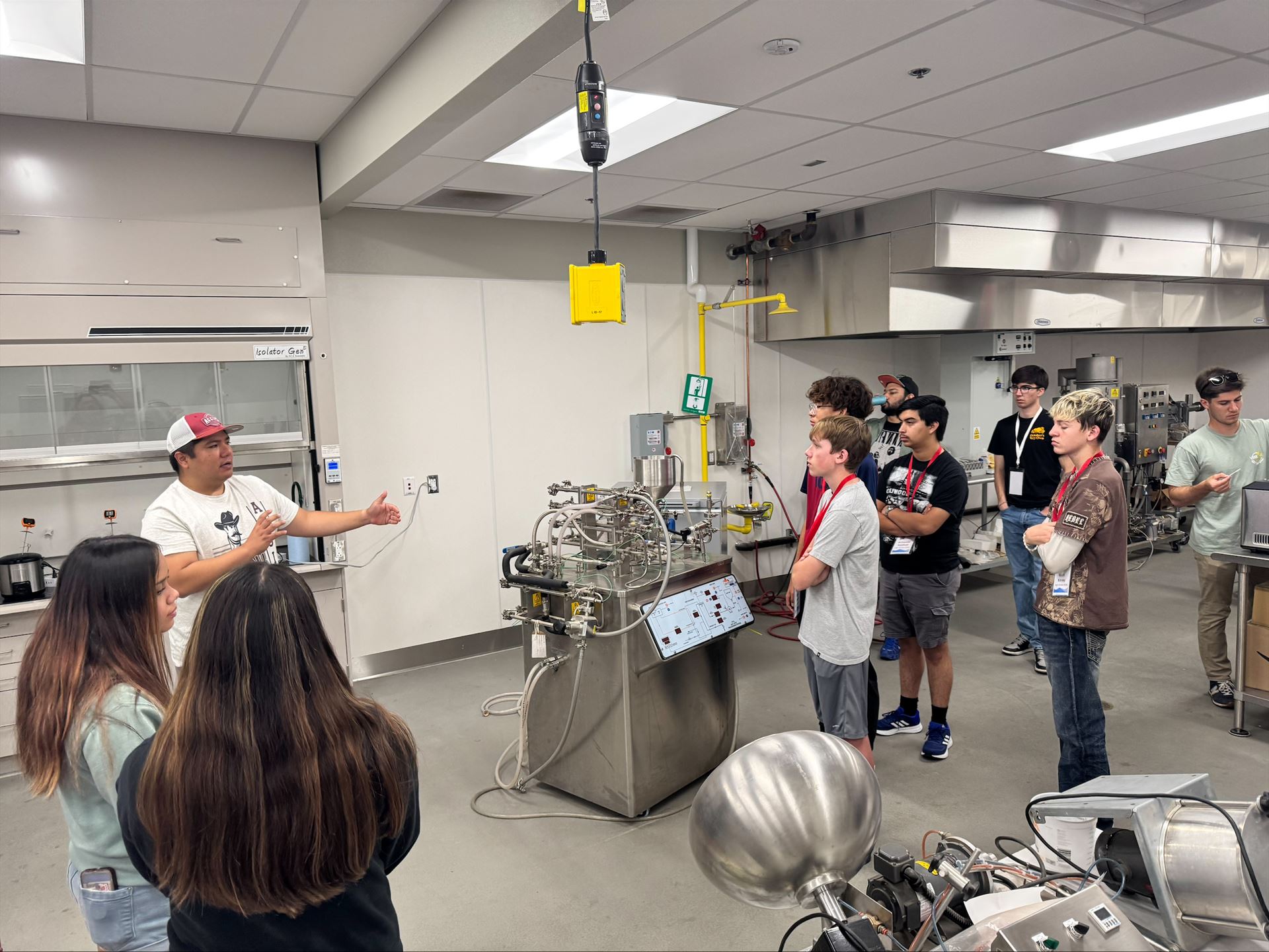 Description High school students attending NM PREP Academy are gathered around a machine in a chemical engineering lab while listening to an instructor
