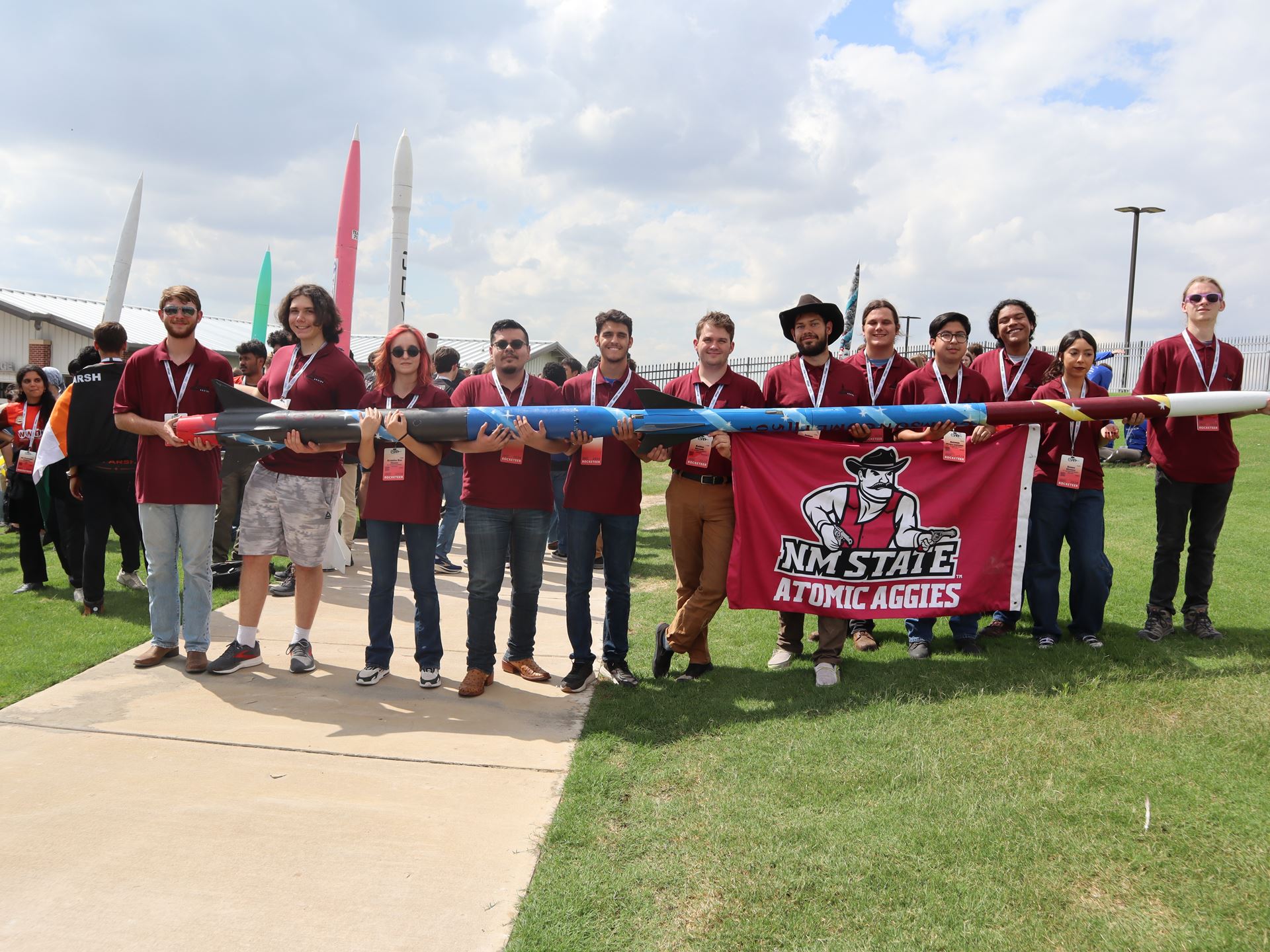 Description The team stands in a line holding the 18 foot rocket they built Under the rocket some members are also holding a banner that reads NM STATE ATOMIC AGGIES