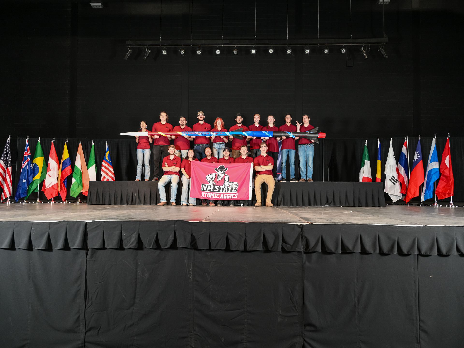 Description The team stands together in two rows The members in the top hold up the rocket the team designed the Crimson Comet II and the members on the bottom hold up a banner that reads NM STATE ATOMIC AGGIES