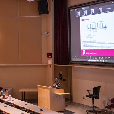 Image description Woman speaking at a podium in a large room with her presentation visible in the background