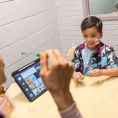 IMAGE DESCRIPTION A boy looks intently at a woman blowing bubbles while a tablet on a table displays learning icons