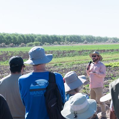 A man in a hat stands outdoors in a field speaking through a microphone to a gathered crowd