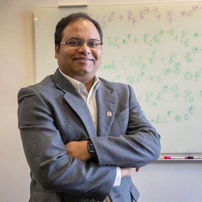 Photo description A man in a suit stands in front of a whiteboard where he has written down code for a project he worked on