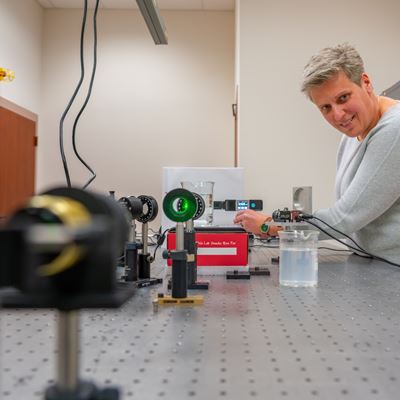 A woman stands in a photonics laboratory where a green laser beam experiment is set up to show its intensity on an optical instrument