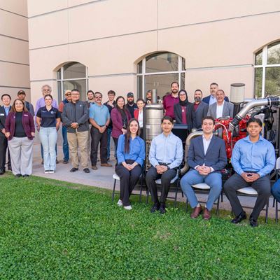 Description A group of people stand and sit next to and behind a large diesel engine that is displayed in a courtyard