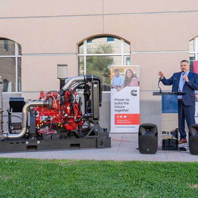 Description A man in a suit stands next to a diesel engine while speaking at an event