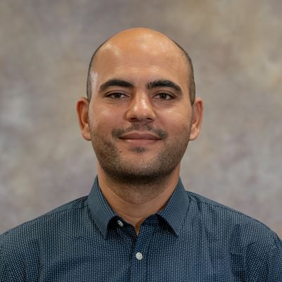 Photo description A man in a blue button down shirt stands for a portrait photo in front of a beige backdrop