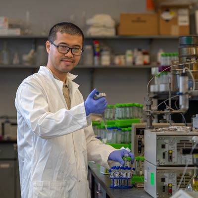 A man in a lab coat stands in a research lab with various instruments used in the field of environmental and civil engineering