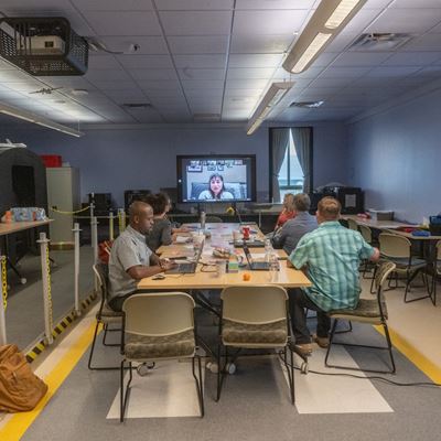 IMAGE DESCRIPTION A group of people seated around a table watching a woman speak on a monitor