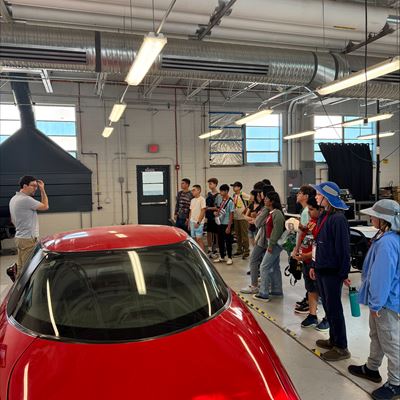 Description Middle school students attending NM PREP Academy are gathered around a red electric car in a mechanical engineering lab while they listen to an instructor