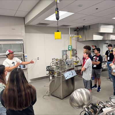 Description High school students attending NM PREP Academy are gathered around a machine in a chemical engineering lab while listening to an instructor