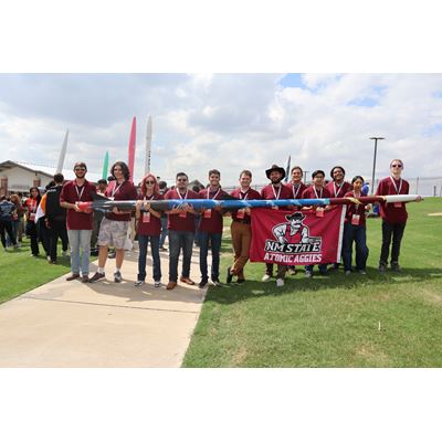 Description The team stands in a line holding the 18 foot rocket they built Under the rocket some members are also holding a banner that reads NM STATE ATOMIC AGGIES