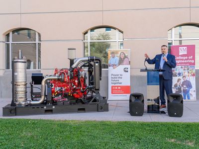 Description A man in a suit stands next to a diesel engine while speaking at an event