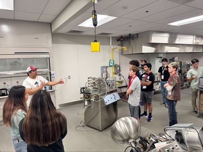 Description High school students attending NM PREP Academy are gathered around a machine in a chemical engineering lab while listening to an instructor
