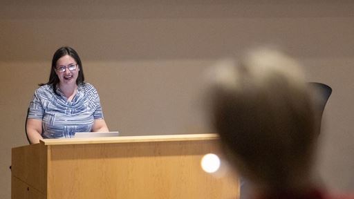 Image description Woman speaking at a podium while attendees listen