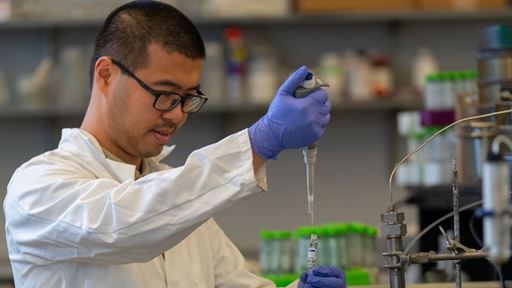 A man in a lab coat holds an instrument used to take liquid samples and move them intro various containers where further testing can be done