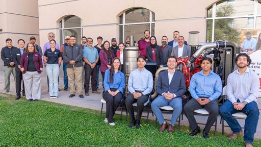Description A group of people stand and sit next to and behind a large diesel engine that is displayed in a courtyard