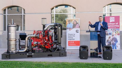 Description A man in a suit stands next to a diesel engine while speaking at an event