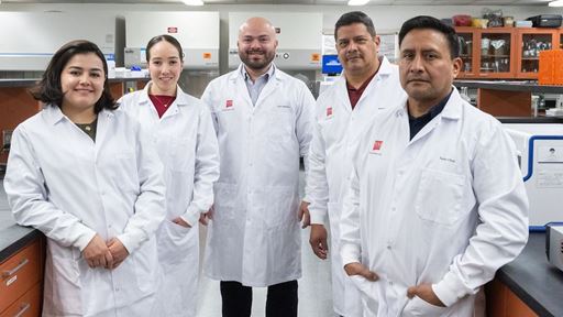 Two women and three man each wearing lab coats standing in the middle of a laboratory