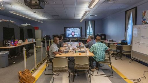 IMAGE DESCRIPTION A group of people seated around a table watching a woman speak on a monitor