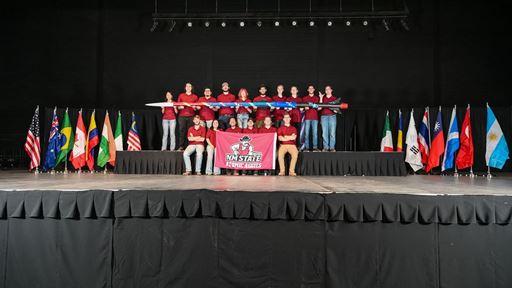 Description The team stands together in two rows The members in the top hold up the rocket the team designed the Crimson Comet II and the members on the bottom hold up a banner that reads NM STATE ATOMIC AGGIES