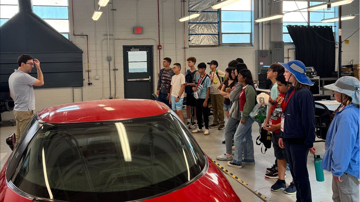 Description Middle school students attending NM PREP Academy are gathered around a red electric car in a mechanical engineering lab while they listen to an instructor