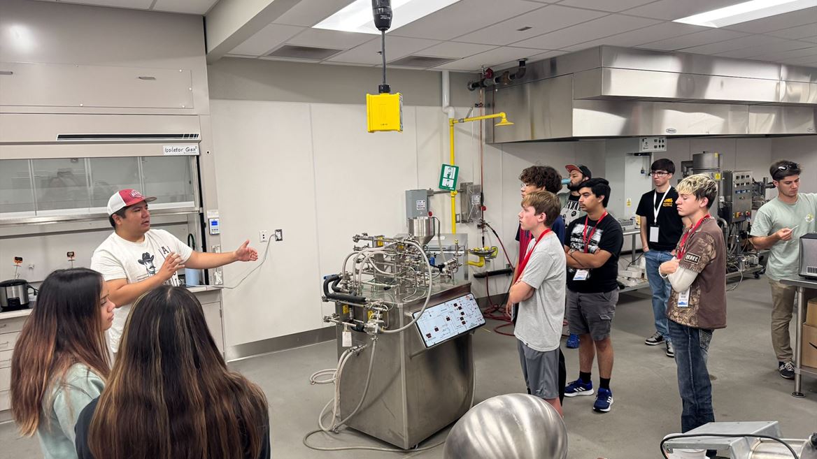 Description High school students attending NM PREP Academy are gathered around a machine in a chemical engineering lab while listening to an instructor
