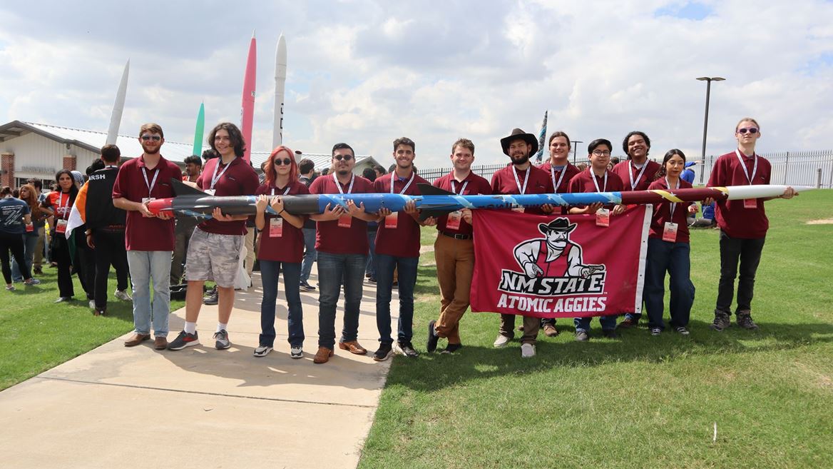 Description The team stands in a line holding the 18 foot rocket they built Under the rocket some members are also holding a banner that reads NM STATE ATOMIC AGGIES